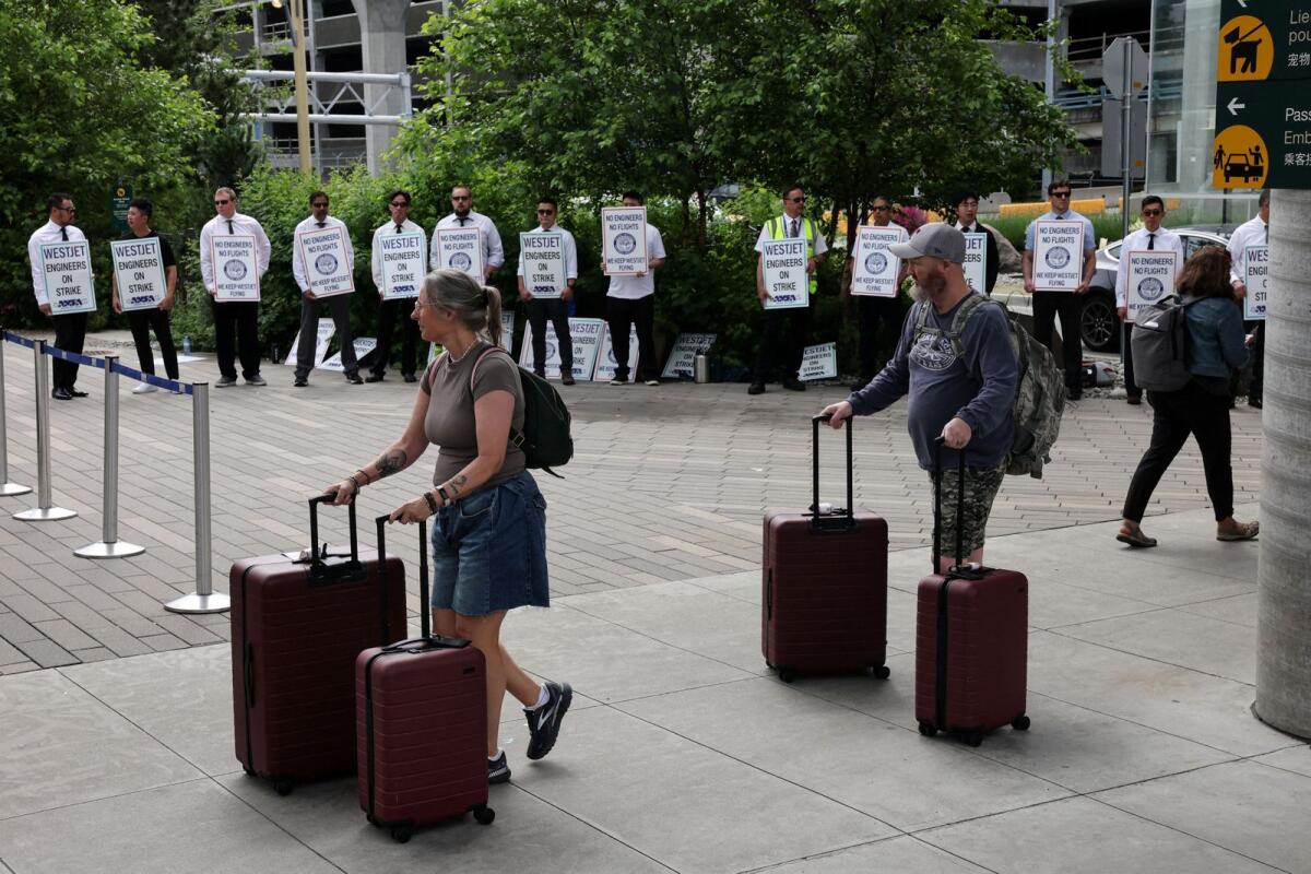 People walk with their luggage as striking aircraft maintenance engineers and technical staff represented by the Aircraft Mechanics Fraternal Association union stand in a picket line against Westjet Airlines at Vancouver International Airport in Richmond, British Columbia, Canada. — Reuters