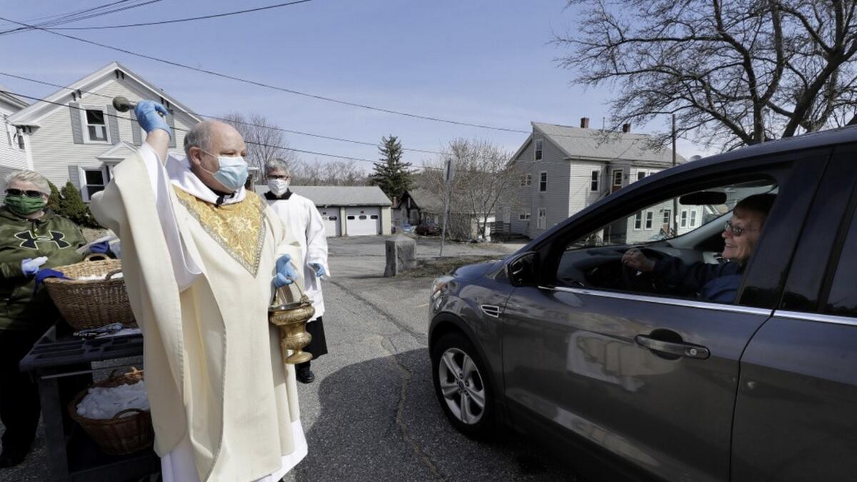 Rev. William Schipper, pastor of Mary, Queen of the Rosary Parish, left, wears a mask and gloves out of concern for the coronavirus as he sprinkles holy water and blesses parishioners who remain in their vehicles in the parking lot of the church, on Easter Sunday. Photo: AP