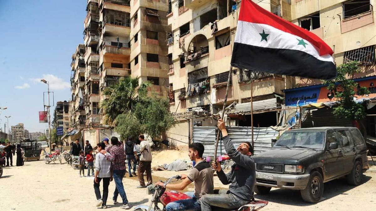 Men waive the Syrian flag as they drive a motorcycle in a street in the Eastern Ghouta town of Douma after Syrian government forces entered the last rebel bastion. — AFP