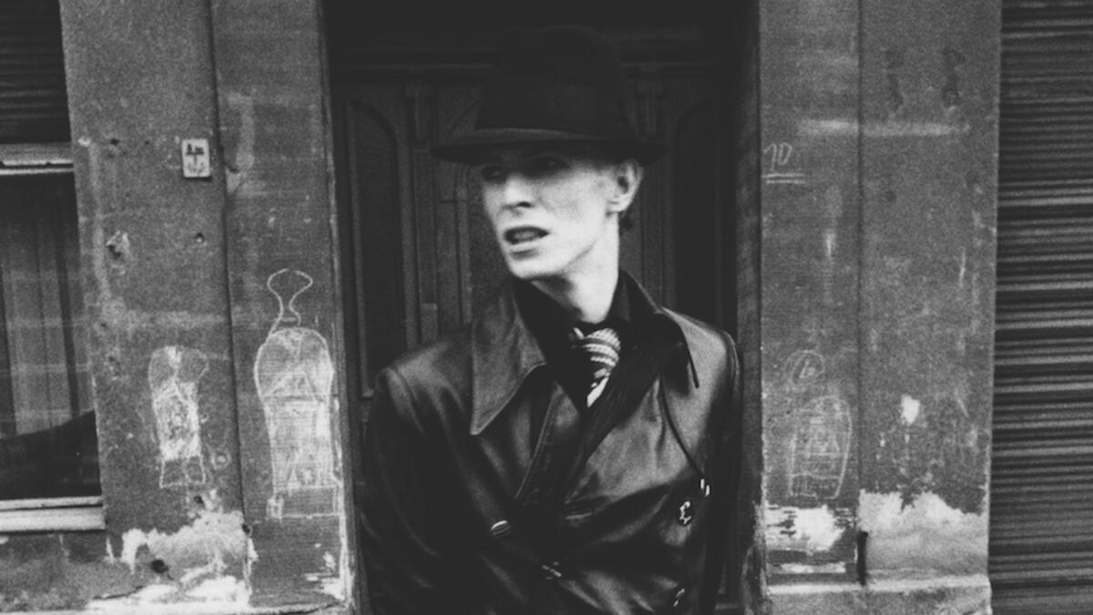 David Bowie dressed in a full length black leather trench coat and hat in East Berlin (1976)