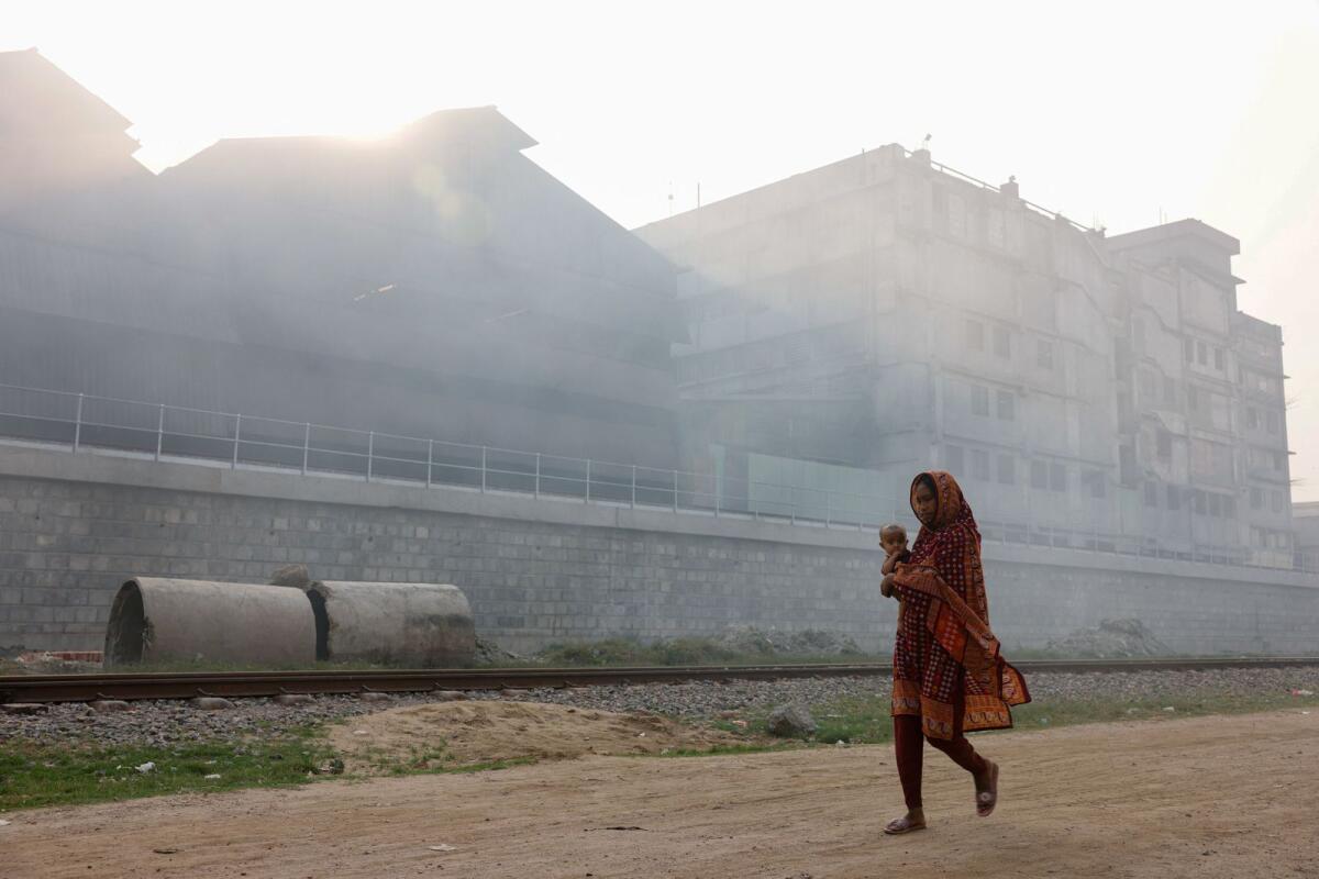 A woman carrying a child passes an industrial area as smoke rises from re-rolling factories at the Shyampur area of Dhaka, Bangladesh, on March 16, 2024. — Reuters