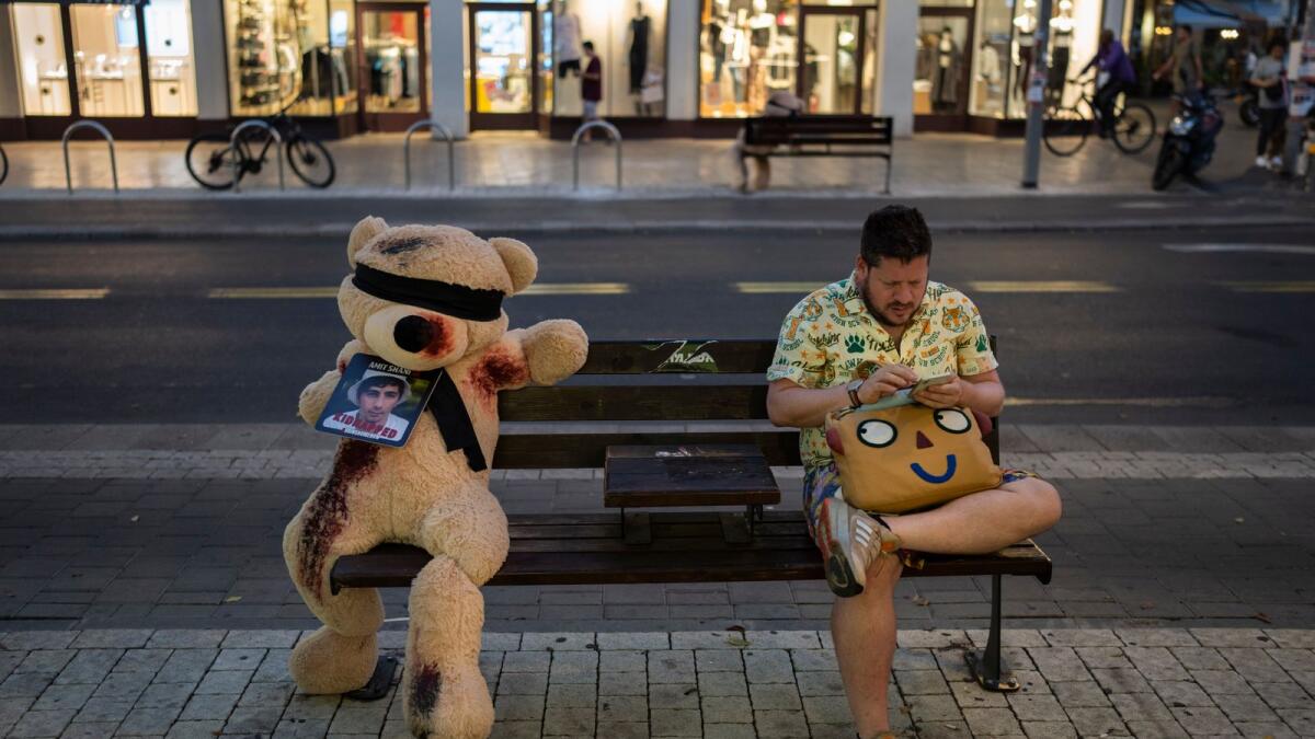A man sits next to an installation of a blindfolded giant teddy bear in Tel Aviv, Israel, Monday, Nov. 6, 2023. The installation is meant to draw attention to over 240 people who were abducted during the Hamas cross-border attack on Oct. 7. (AP Photo/Bernat Armangue)