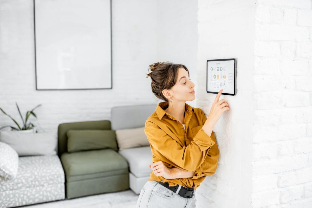 Woman controlling home with a digital touch screen panel installed on the wall in the living room. The luxury real estate market will be progressively integrating eco-friendly features and designs, in keeping with global developments in sustainable living.